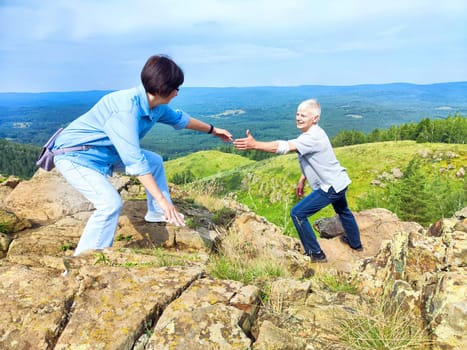 One woman reaches out to assist another as they navigate a rocky slope surrounded by expansive greenery and hills.