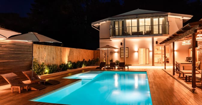 Modern house with illuminated swimming pool and spacious wooden deck at night. Luxury outdoor space designed for relaxation and entertainment