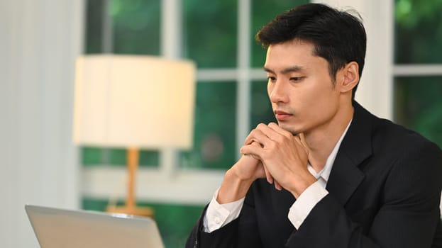 Thoughtful millennial entrepreneur with his hands clasped under his chin working on laptop.
