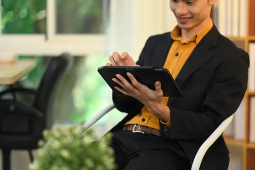 Handsome asian businessman in formal suit working on digital tablet at modern workplace.