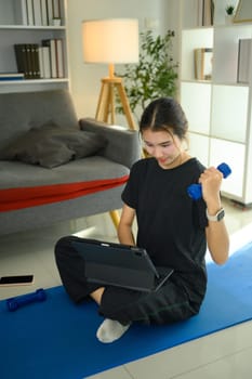 Young asian woman lifting a light dumbbell and using digital tablet on yoga mat.