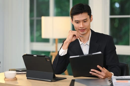 Handsome businessman in formal suit working on digital tablet at modern workplace.