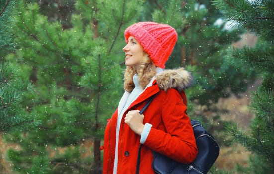 Happy young woman looking away with backpack, enjoys snow, warm weather against Christmas tree background, joyful girl in red hat standing in snowy forest with snowflakes