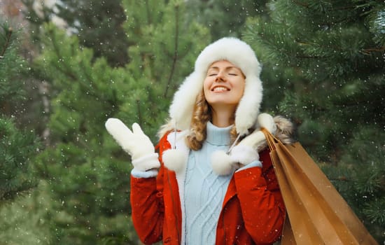 Happy smiling young woman with shopping bag in winter against Christmas tree background with snow, joyful girl in white hat standing in snowy forest