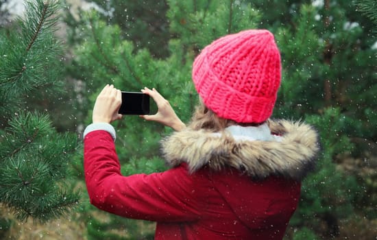 Young woman taking picture on phone Christmas tree in winter park, girl with smartphone outdoors, wearing red jacket, hat, standing in snowy forest with snowflakes, nature background