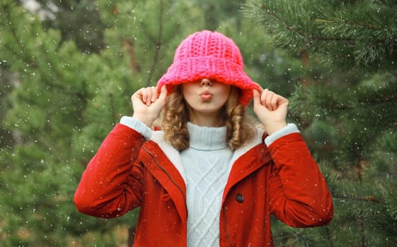 Happy cheerful young woman having fun enjoys snow, warm weather in winter park against Christmas tree, joyful girl in hat standing in forest with snowflakes, festive background