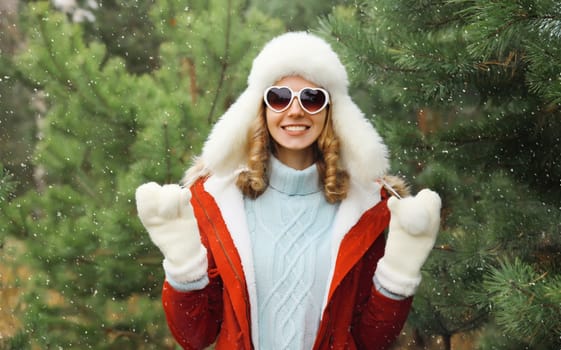 Happy smiling young woman having fun enjoys snow, warm weather in winter park against Christmas tree, joyful girl in hat, glasses standing in forest with snowflakes, festive background