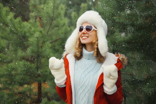 Happy smiling young woman having fun enjoys snow, warm weather in winter park against Christmas tree, joyful girl in hat, glasses standing in forest with snowflakes, festive background