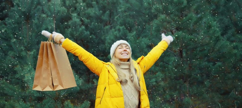 Happy smiling young woman with shopping bag in winter against Christmas tree, joyful girl in yellow jacket, hat standing in snowy forest
