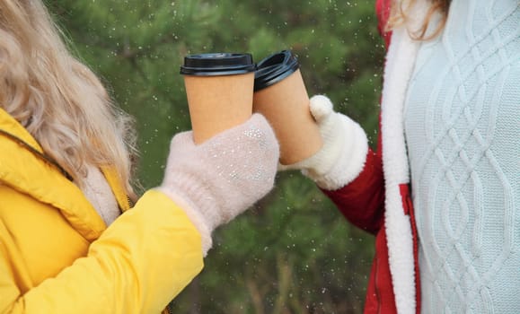 Close up two women friends, female hands holding cups of hot coffee drink against Christmas trees in winter forest