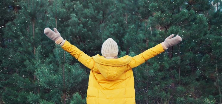 Back view, winter day, happy young woman enjoys snow, warm weather against Christmas tree, joyful girl in hat, yellow jacket raising her hands up in snowy green forest with snowflakes