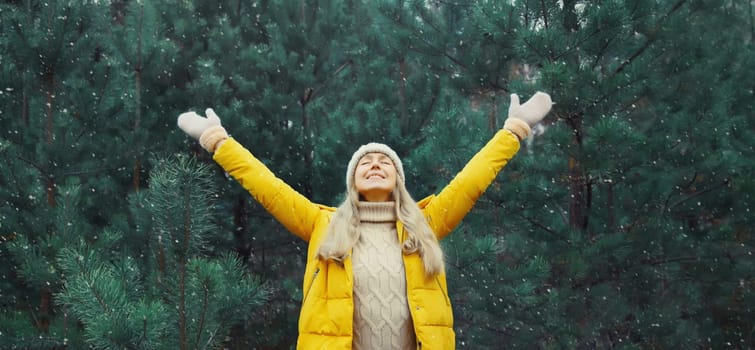Winter day, happy young woman enjoys snow, warm weather against Christmas tree, joyful girl in hat, yellow jacket raising her hands up in snowy green forest with snowflakes