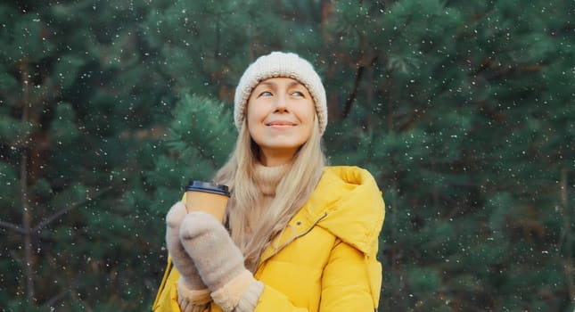 Winter day, happy smiling mature woman holding cup of coffee drink enjoys snow, warm weather against Christmas tree, wearing yellow jacket, hat, standing in snowy forest with snowflakes