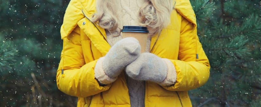 Close up female hand holding cup of hot coffee drink against Christmas trees in winter forest, wearing yellow jacket, mittens