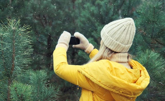 Young woman taking picture on phone Christmas tree in winter park, girl with smartphone outdoors, wearing yellow jacket, hat, standing in snowy forest with snowflakes, nature background