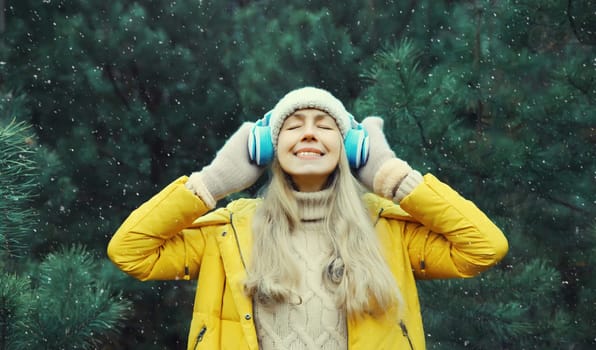 Portrait of happy modern young woman enjoying listening to music with headphones in hat, bright yellow jacket in winter forest against Christmas tree background with snow