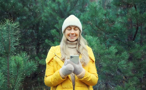 Happy smiling young woman with phone in winter against Christmas tree, joyful girl using smartphone in white hat, yellow jacket standing in snowy forest