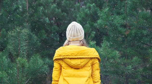 Back view, winter day, happy young woman enjoys snow, warm weather against Christmas tree, joyful girl in hat, yellow jacket in snowy green forest with snowflakes