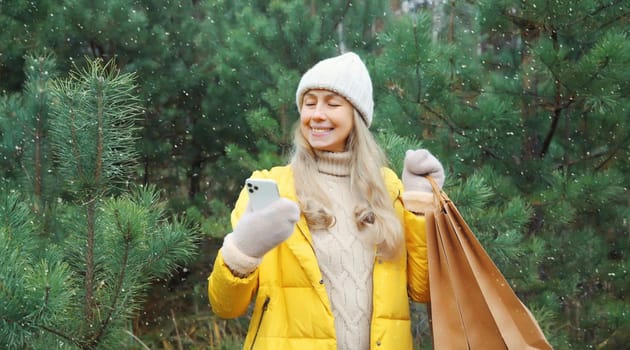 Happy smiling mature woman with phone, shopping bag in winter against Christmas tree, joyful girl using smartphone in white hat, yellow jacket standing in snowy forest