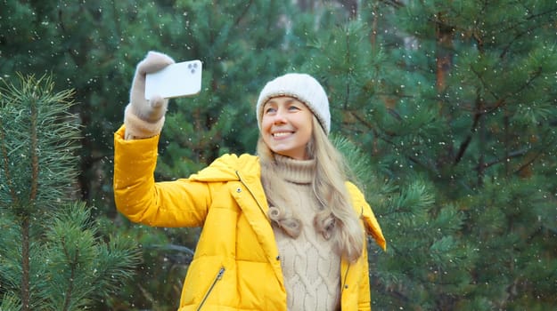 Portrait of happy smiling young woman taking selfie with smartphone in winter park against Christmas tree, joyful girl using phone in hat, yellow jacket in forest