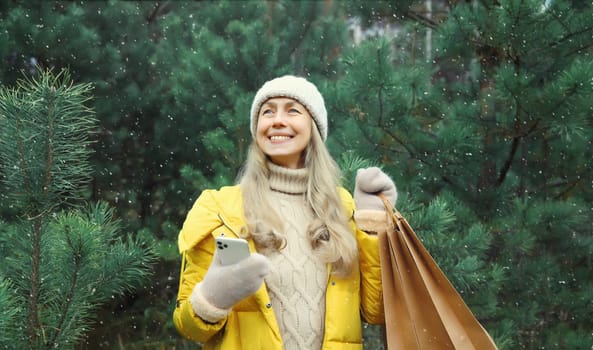 Happy smiling mature woman with phone, shopping bag in winter against Christmas tree, joyful girl using smartphone in white hat, yellow jacket standing in snowy forest