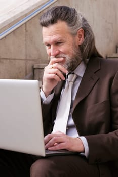 pensive mature male entrepreneur in elegant suit siting on urban stairway and working online on laptop in city, concept of business and middle age man lifestyle