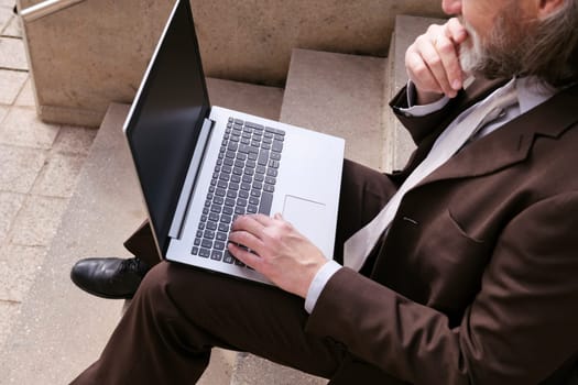 crop of unrecognizable male entrepreneur in formal clothes sitting on urban stairway and working online on laptop in city, concept of business and remote work