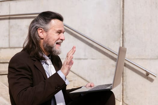 positive middle age male entrepreneur in elegant suit siting on stairway and waving hand saying hello while having online video meeting via laptop, concept of business and middle age man lifestyle