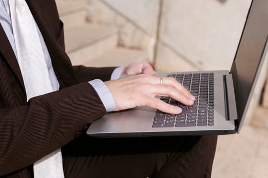 close up of the hands of an unrecognizable male entrepreneur in formal clothes sitting on urban stairway and working online on laptop in city, concept of business and remote work