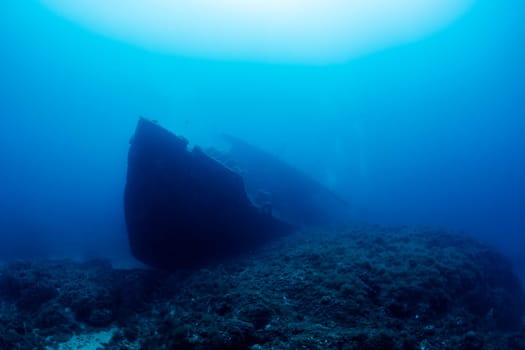 wreck of an old ruined ship on ocean rocky bottom covered with moss and algae in deep blue sea with clear water and light glare, concept of underwater adventure and seascape