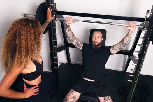 concentrated sportsman with tattoos lifting barbell with heavy weights during functional workout with female instructor in gym, concept of fitness and active lifestyle