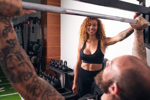 unrecognizable sportsman with tattoos lifting barbell with heavy weights during functional workout with female instructor in gym, concept of fitness and active lifestyle