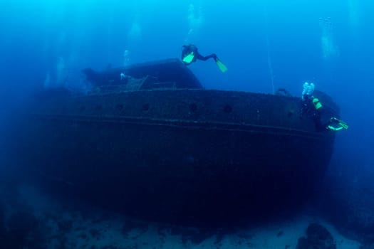 two scuba divers diving near of the wreck of an old ruined ship on the bottom of deep blue sea with clear water and light glare, concept of underwater adventure and seascape