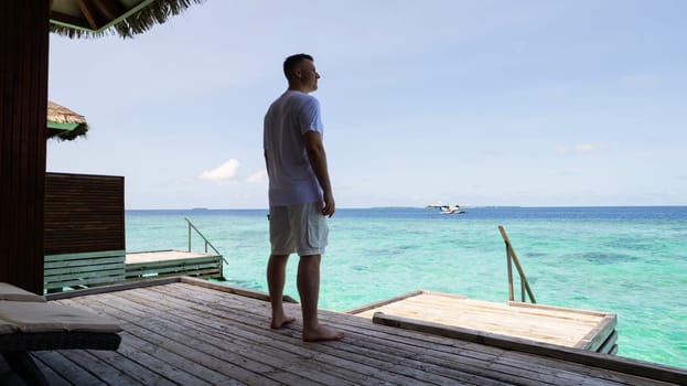 A guy in white clothes admires the turquoise ocean. Vacation in the Maldives. He looks into the future and enjoys it. Small waves. Descent into the water. Wooden villa. Bungalows on the water