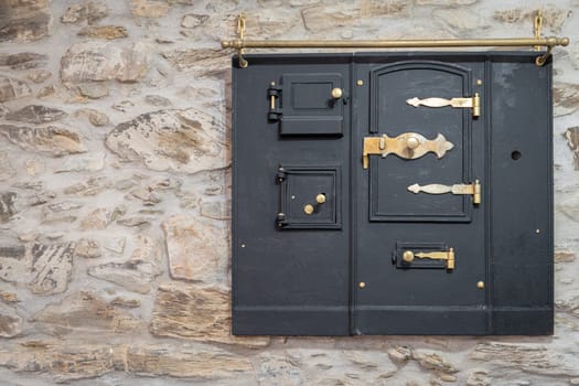A rustic black metal door with ornate brass hinges and locks on a textured stone wall blends vintage and industrial styles.