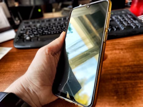 A person holds a cell phone with a glossy screen while it charges on a wooden surface in an office environment.