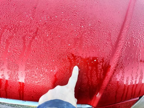 A person points at noticeable damage on a wet red car in a parking lot during rain.