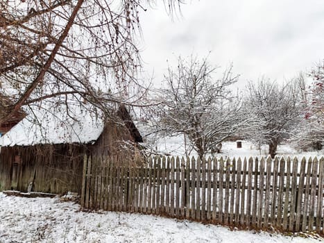 A tranquil winter scene showcases a rustic house beside snow-dusted trees and a charming wooden fence in a rural area.