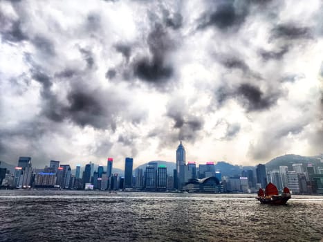 Hong Kong - April 05, 2024: A vibrant ship navigates through the waters of Victoria Bay, with Hong Kong's skyline looming in the backdrop beneath a moody sky filled with clouds. The scene captures urban life and natural beauty.