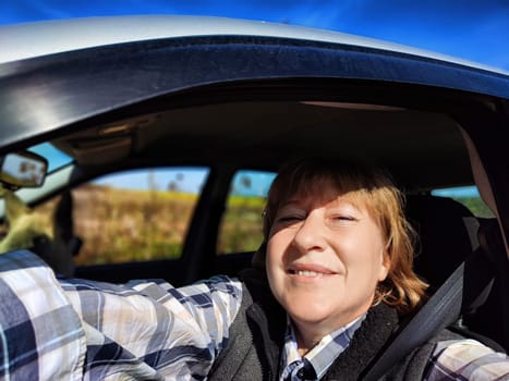 A middle-aged woman smiles happily as she takes a selfie in her car while driving through a picturesque countryside landscape.