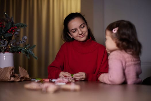 mother and her little child girl together wrapping Christmas gifts at home in the evening, preparing presents for family.