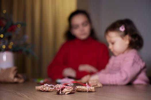 Christmas gifts on table and mother and little child girl wrapping Christmas gifts at background, focus on gifts