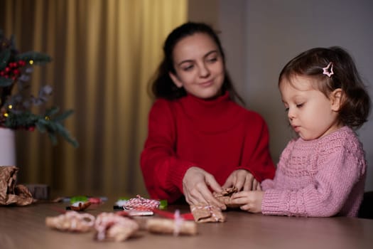 mother and her little child girl together wrapping Christmas gifts at home in the evening, preparing presents for family.