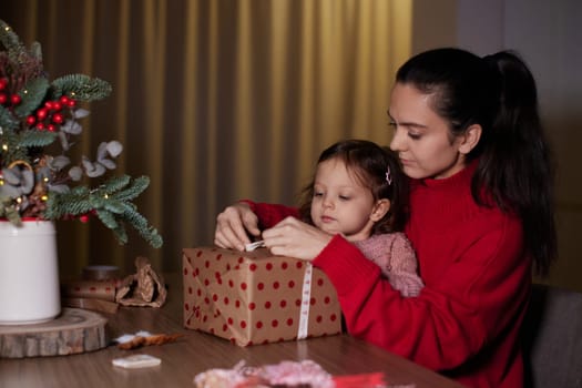 mother and her little child girl together wrapping Christmas gifts at home in the evening, preparing presents for family.