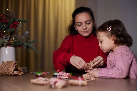 mother and her little child girl together wrapping Christmas gifts at home in the evening, preparing presents for family.