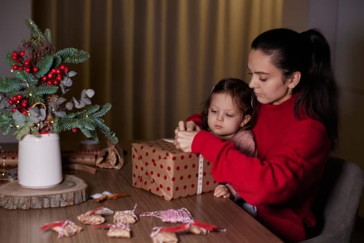 mother and her little child girl together wrapping Christmas gifts at home in the evening, preparing presents for family.