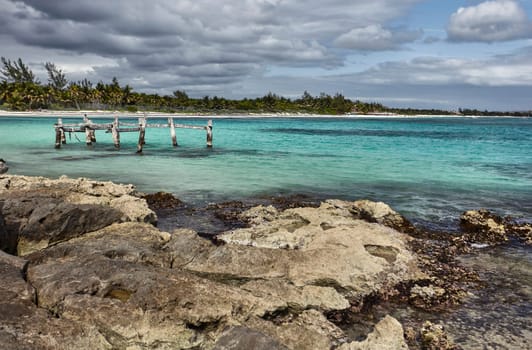 Wooden poles of an old pier submerged in the turquoise waters of xpu-ha beach, with rocks in the foreground and a cloudy sky