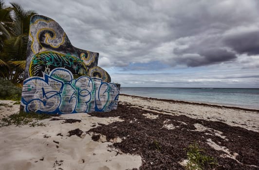 Vibrant graffiti covers a concrete slab on the sandy shores of xpu-ha beach in mexico's riviera maya, with a cloudy sky overhead