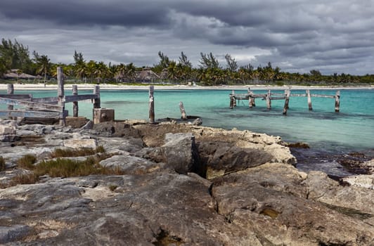 Decaying wooden pier stretches into the turquoise waters of the caribbean sea, under a dramatic cloudy sky in xpu-ha, mexico
