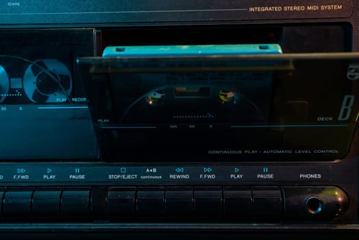 Close-up of a vintage stereo cassette deck playing a turquoise audio cassette, illuminated by dark ambient light, evoking nostalgia for the golden age of analog music
